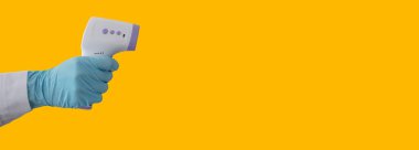 Koruyucu eldiven giyen ve kızılötesi termometre kullanmaya hazır bir doktorun eli, sarı arka planda izole edilmiş, salgın virüsü salgını kavramı.