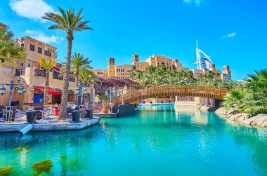 Park area of Souk Madinat Jumeirah market complex with canal, pedestrian bridge, lush greenery, Fort Island, cafes and Burj Al Arab in background, Dubai, UAE