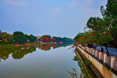 The wide Ping river with clear mirror surface, lush green parks along the banks and blooming flame trees, Chiang Mai, Thailand