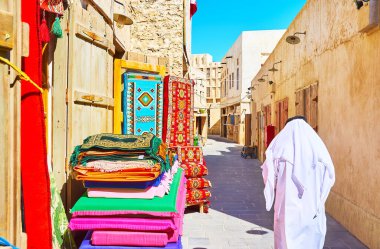 Souq Waqif 'in dar sokaklarındaki halı mağazasında, yerli halk geleneksel giysiler içinde yol boyunca yürüyorlar - keffiyeh (türban) ve bulaşık bezi (bornoz), Doha, Katar