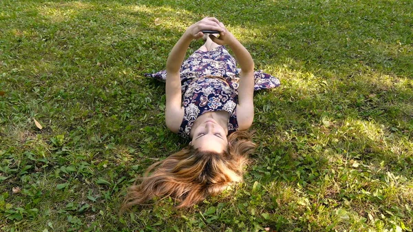 A young girl in a dress lies on the green grass with a phone in her hands. The girl lies on her back in the summer park. — Stock Photo, Image