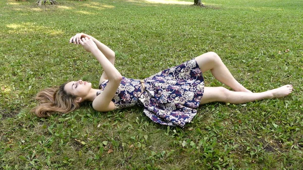 Uma jovem em um vestido jaz na grama verde com um telefone em suas mãos. — Fotografia de Stock