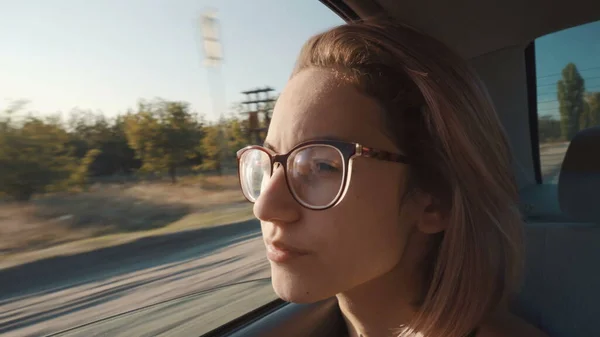 Ένα κορίτσι με γυαλιά κάθεται στο πίσω κάθισμα και κοιτάει έξω από το ανοιχτό παράθυρο.. — Φωτογραφία Αρχείου