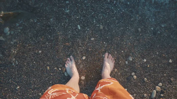 Un joven está parado en una playa de piedra y la ola cubre sus piernas. Vista superior de las piernas. — Foto de Stock