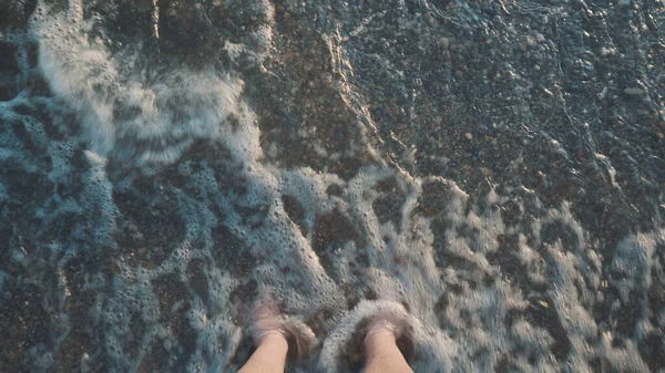 Um jovem está em uma praia de pedra e a onda cobre suas pernas. Vista superior das pernas. — Fotografia de Stock