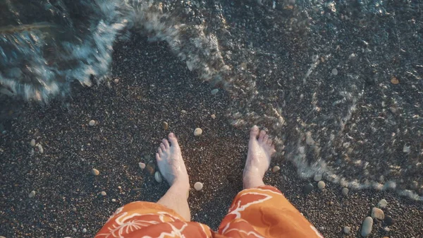 Ein junger Mann steht an einem steinernen Strand und die Welle bedeckt seine Beine. Blick von oben auf die Beine. — Stockfoto