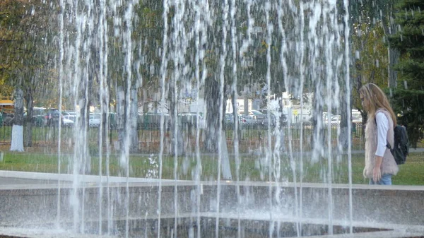Poblíž fontány kráčí mladá žena. Pohled skrz fontánu — Stock fotografie