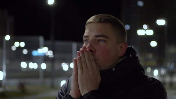 Молодой человек стоит на холоде вечером на улице и согревает руки своим дыханием. — стоковое видео