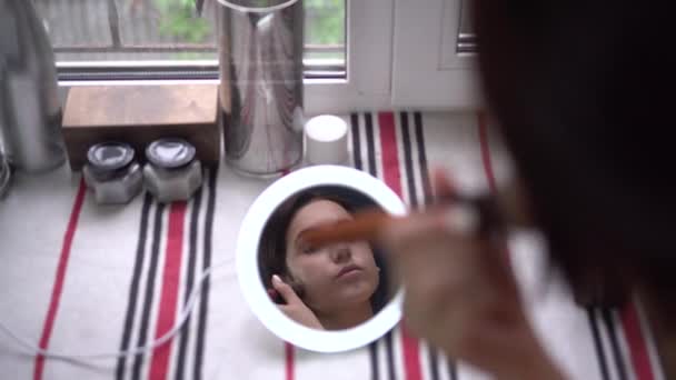 En ung kvinna framför fönstret tittar in i en liten belyst spegel och applicerar makeup med en borste. Utsikt över flickan genom spegeln. — Stockvideo