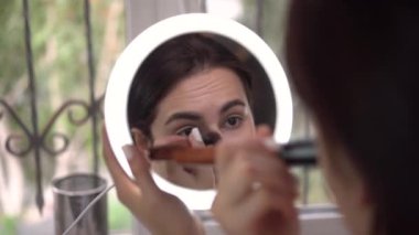 Pencerenin önündeki genç bir kadın küçük ışıklı bir aynaya bakar ve bir fırçayla makyaj yapar. Aynadan kıza bak..