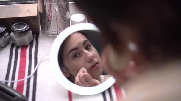 Uma jovem mulher na frente de uma janela olha para um pequeno espelho com luz e aplica maquiagem com uma esponja. Vista da menina através do espelho. — Vídeo de Stock