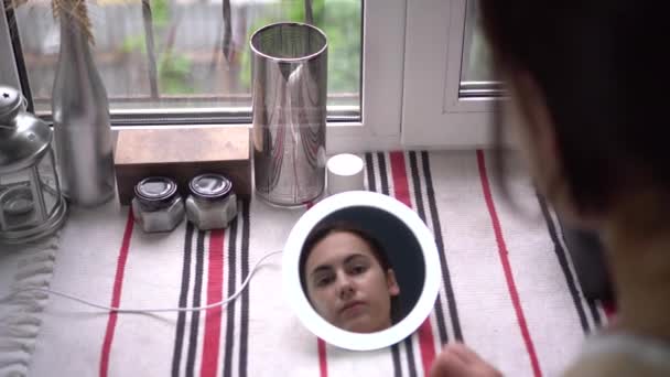 Μια νεαρή γυναίκα ανάβει το φως σε ένα μικρό καθρέφτη και απλώνει το μακιγιάζ της με ένα σφουγγάρι. Άποψη του κοριτσιού μέσα από τον καθρέφτη. — Αρχείο Βίντεο