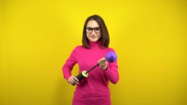 Genç bir kadın, sarı arka planda pompası olan mor bir balonu şişiriyor. Pembe balıkçı yaka ve gözlüklü kız..