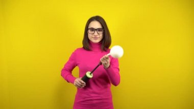 Genç bir kadın sarı arka planda pompası olan beyaz bir balonu şişiriyor. Pembe balıkçı yaka ve gözlüklü kız..