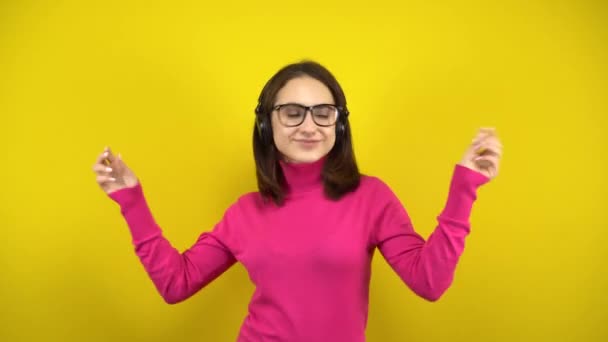 Молода жінка танцює з навушниками на жовтому тлі. Дівчина в рожевому горлику і окулярах слухає музику . — стокове відео