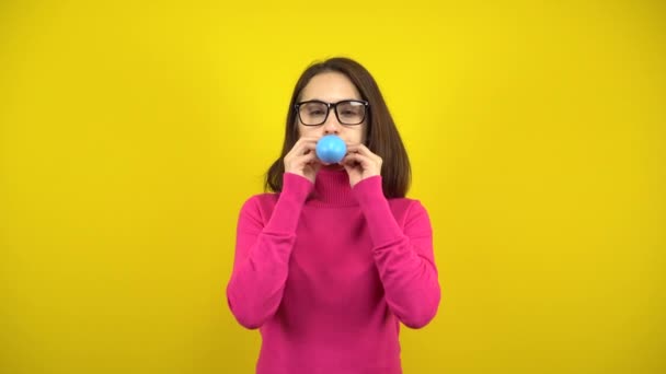 En ung kvinna blåser upp en blå ballong med munnen på en gul bakgrund. Flicka i rosa polotröja och glasögon. — Stockvideo