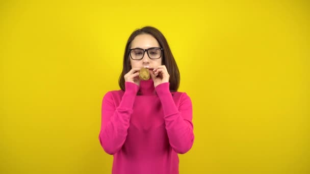 A young woman inflates a gold balloon with her mouth on a yellow background. Girl in a pink turtleneck and glasses. — Stock Video