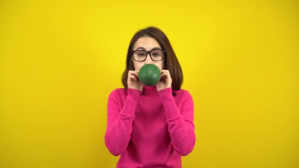 Uma jovem infla um balão verde com a boca em um fundo amarelo. Menina em uma gola alta rosa e óculos. — Vídeo de Stock