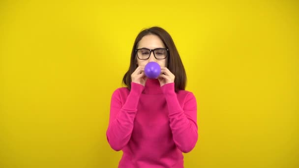 Uma jovem infla um balão roxo com a boca em um fundo amarelo. Menina em uma gola alta rosa e óculos. — Vídeo de Stock