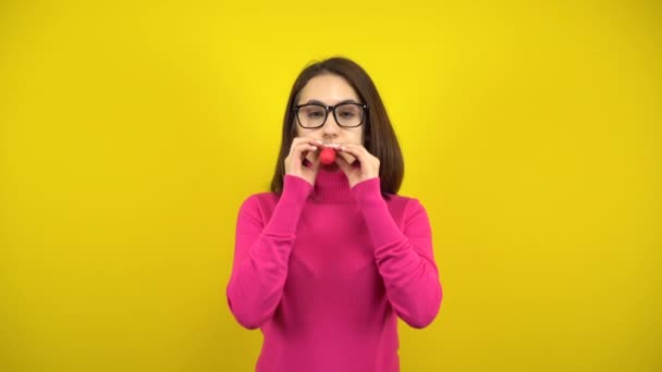 En ung kvinna blåser upp en röd ballong med munnen på en gul bakgrund. Flicka i rosa polotröja och glasögon. — Stockvideo