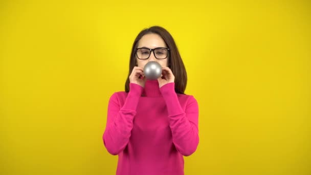 En ung kvinna blåser upp en silverballong med munnen på en gul bakgrund. Flicka i rosa polotröja och glasögon. — Stockvideo