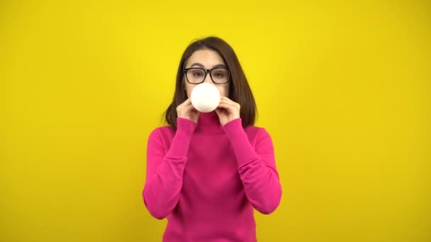 Mladá žena nafoukne bílý balón s pusou na žlutém pozadí. Dívka v růžovém roláku a brýle. — Stock video