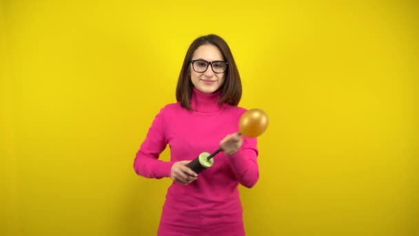 Eine junge Frau bläst einen goldenen Luftballon mit einer Pumpe auf gelbem Grund auf. Mädchen mit rosa Rollkragen und Brille. — Stockvideo