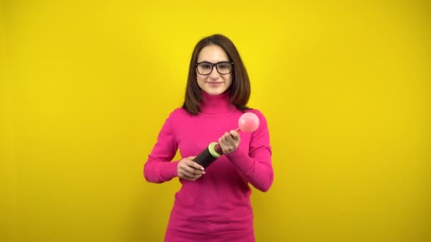 En ung kvinna blåser upp en rosa ballong med en pump på en gul bakgrund. Flicka i rosa polotröja och glasögon. — Stockvideo