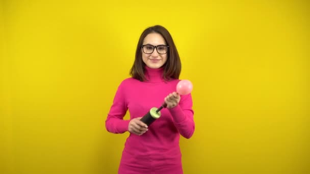 En ung kvinna blåser upp en rosa ballong med en pump och den spricker på en gul bakgrund. Flicka i rosa polotröja och glasögon. — Stockvideo