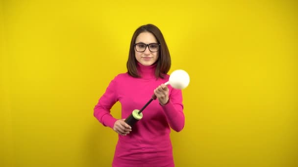 Een jonge vrouw blaast een witte ballon op met een pomp op een gele achtergrond. Meisje in een roze coltrui en bril. — Stockvideo