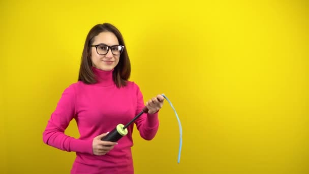 Una joven infla un globo azul largo con una bomba sobre un fondo amarillo. Chica en un cuello alto rosa y gafas. — Vídeos de Stock