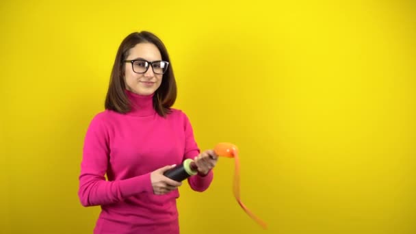 En ung kvinna blåser upp en lång orange ballong med en pump på en gul bakgrund. Flicka i rosa polotröja och glasögon. — Stockvideo