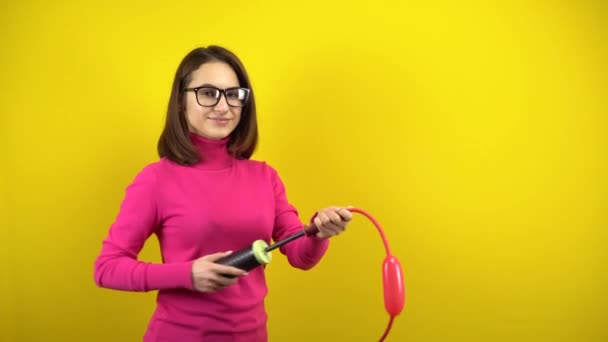 Uma jovem infla um longo balão vermelho com uma bomba sobre um fundo amarelo. Menina em uma gola alta rosa e óculos. — Vídeo de Stock