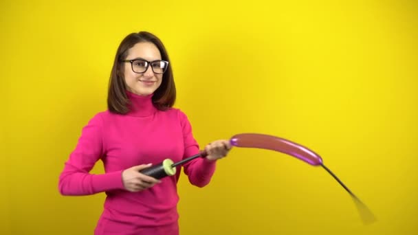 En ung kvinna blåser upp en lång lila ballong med en pump och den brister på en gul bakgrund. Flicka i rosa polotröja och glasögon. — Stockvideo