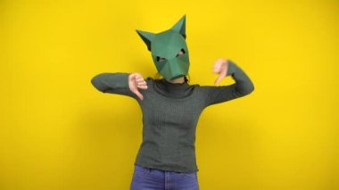 Kartondan çakal maskeli genç bir kadın sarı arka planda baş parmaklı hoşlanmadığını gösteriyor. Yeşil kazaklı ve maskeli kadın..