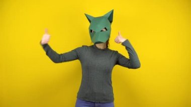 Kartondan çakal maskeli genç bir kadın sarı arka planda baş parmağını gösteriyor. Yeşil kazaklı ve maskeli kadın..