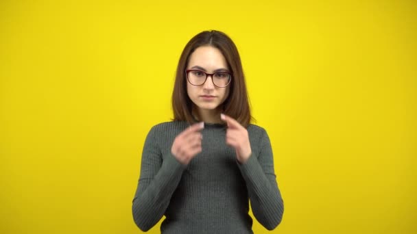 Uma jovem faz um sorriso triste em seu rosto com os dedos em um fundo amarelo. Mulher com óculos e uma camisola verde. — Vídeo de Stock