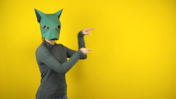 Una giovane donna con una maschera di sciacallo di cartone indica lo spazio vuoto con le dita indice su uno sfondo giallo. Donna in maglione verde e maschera. — Video Stock