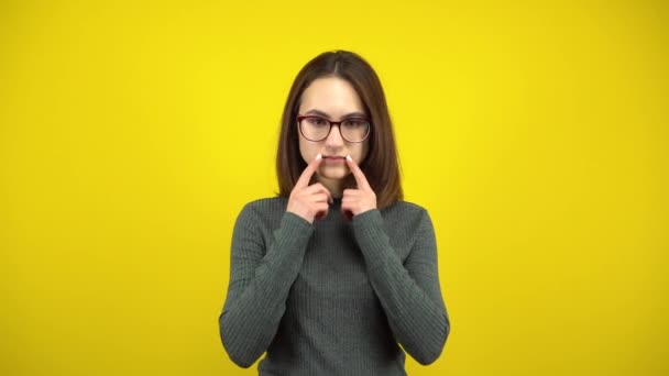 Eine junge Frau macht ein fröhliches Lächeln auf ihrem Gesicht, ihre Finger auf gelbem Hintergrund. Frau mit Brille und grünem Pullover. — Stockvideo