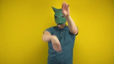 Kartondan bir çakal maskeli genç adam sarı arka planda dans ediyor. Yeşil polo ve maskeli bir adam..