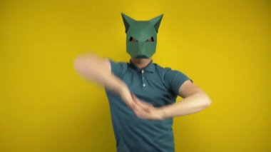 Kartondan bir çakal maskeli genç adam sarı arka planda dans ediyor. Yeşil polo ve maskeli bir adam..