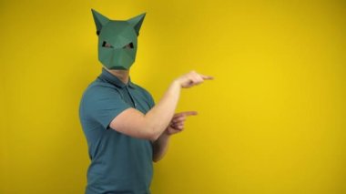 Karton maskeli genç bir adam sarı arka planda işaret parmaklarıyla boş alanı işaret ediyor. Yeşil polo ve maskeli bir adam..