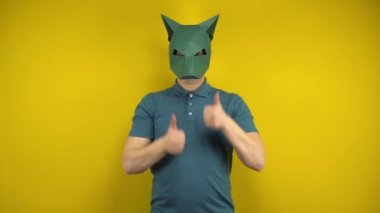 Kartondan çakal maskeli genç bir adam sarı arka planda baş parmağını gösteriyor. Yeşil polo ve maskeli bir adam..