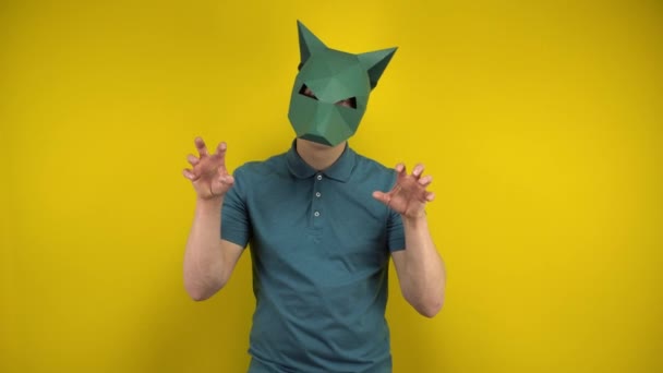 Un joven con una máscara de chacal de cartón representa a un animal y mira a la cámara sobre un fondo amarillo. Hombre en un polo verde y máscara. — Vídeos de Stock