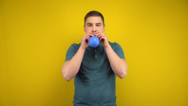 Un giovane gonfia un palloncino blu con la bocca su uno sfondo giallo. Uomo in polo verde. — Video Stock