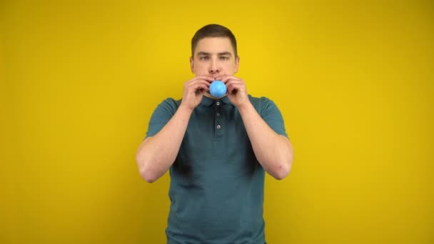 En ung man blåser upp en blå ballong med munnen på en gul bakgrund. Man i grön polo. — Stockvideo
