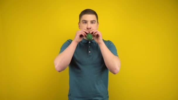 En ung man blåser upp en grön ballong med munnen på en gul bakgrund. Man i grön polo. — Stockvideo