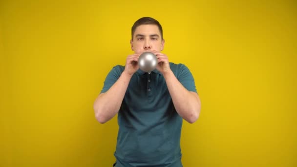 Un giovane gonfia un palloncino d'argento con la bocca su uno sfondo giallo. Uomo in polo verde. — Video Stock