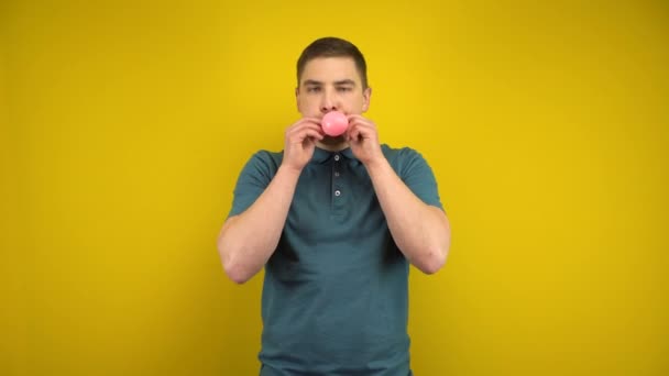 Молодой человек надувает розовый шарик ртом на жёлтом фоне. Человек в зелёном поло. — стоковое видео
