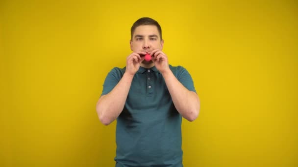 Un joven infla un globo rojo con la boca sobre un fondo amarillo. Hombre en un polo verde. — Vídeo de stock